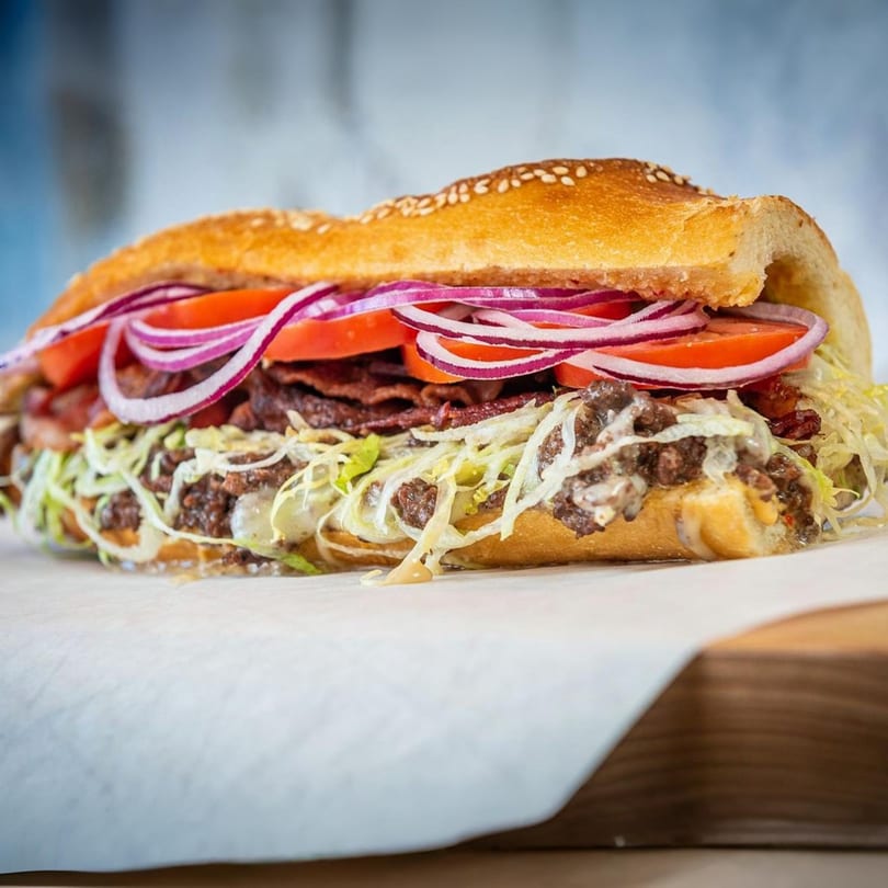 A large, loaded sub sandwich with meat, cheese, lettuce, tomatoes, and red onions on a cutting board.